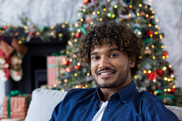 Christmas portrait of young happy man, Hispanic man smiling and looking at camera, happy and celebrating Christmas sitting on sofa in living room near decorated Christmas tree.