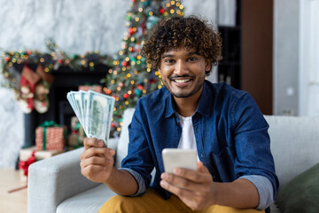 Portrait of joyful man on Christmas at home, hispanic man looking at camera sitting on sofa near...