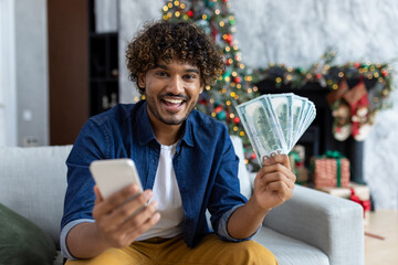 Portrait of joyful man on Christmas at home, hispanic man looking at camera sitting on sofa near...