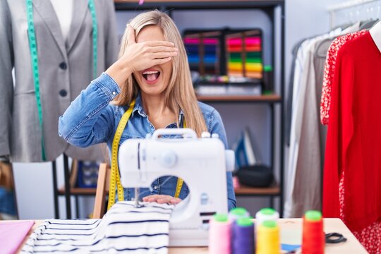 Blonde woman dressmaker designer using sew machine smiling and laughing with hand on face covering eyes for surprise. blind concept.