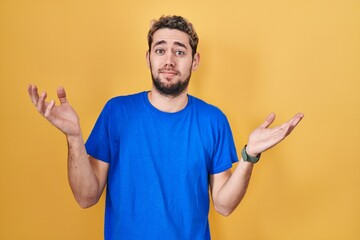 Hispanic man with beard standing over yellow background clueless and confused expression with arms and hands raised. doubt concept.