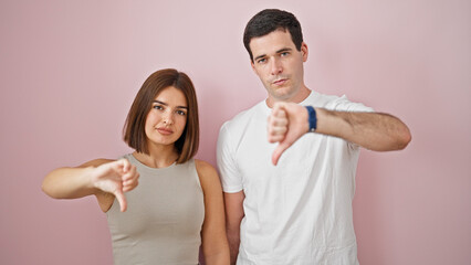 Beautiful couple standing together with serious face doing thumb down gesture over isolated pink background