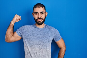 Middle east man with beard standing over blue background strong person showing arm muscle, confident and proud of power