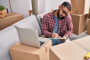 Young hispanic man talking on smartphone writing on document at new home