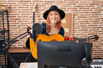Young caucasian woman playing classic guitar at music studio celebrating achievement with happy smile and winner expression with raised hand