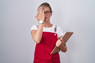 Young blonde woman wearing waiter uniform holding clipboard covering one eye with hand, confident smile on face and surprise emotion.