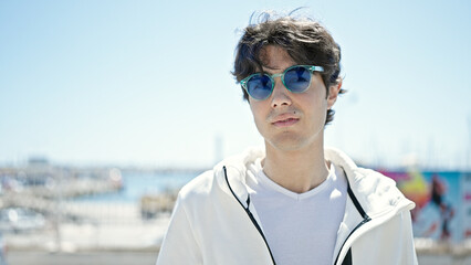 Young hispanic man standing with serious expression wearing sunglasses at seaside