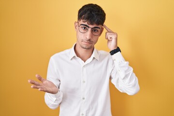 Young hispanic man standing over yellow background confused and annoyed with open palm showing copy space and pointing finger to forehead. think about it.