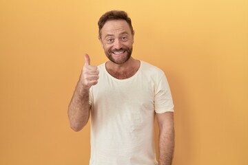 Middle age man with beard standing over yellow background doing happy thumbs up gesture with hand. approving expression looking at the camera showing success.