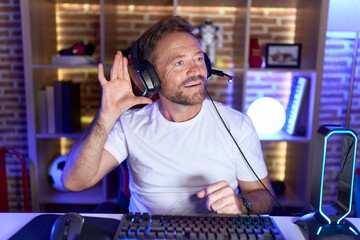 Middle age man with beard playing video games wearing headphones smiling with hand over ear listening an hearing to rumor or gossip. deafness concept.