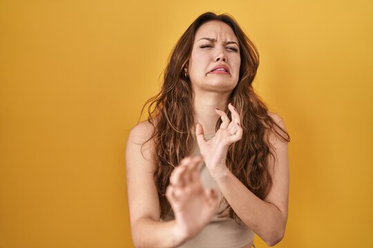 Young hispanic woman standing over yellow background disgusted expression, displeased and fearful doing disgust face because aversion reaction. with hands raised