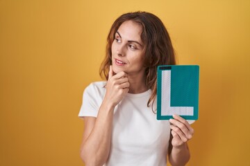 Beautiful brunette woman holding l sign for new driver serious face thinking about question with hand on chin, thoughtful about confusing idea