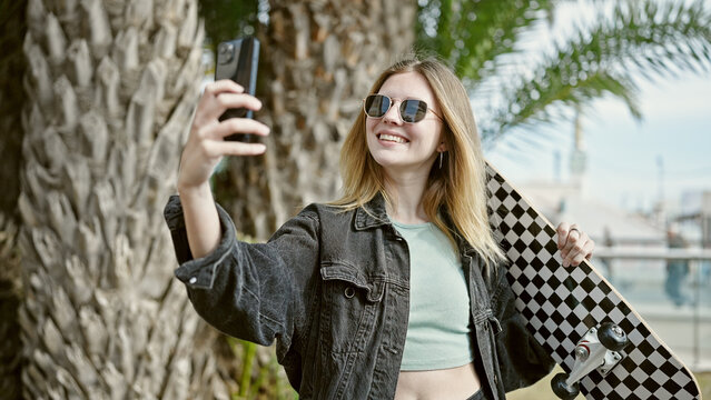 Young blonde woman holding skate make selfie by smartphone smiling at park