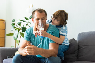 Happy child boy treats his dad at home. Medical procedures. Inhaler. Respiratory medicine. Bronchitis, asthmatic health equipment. Concept of home treatment - obrazy, fototapety, plakaty