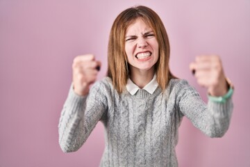 Beautiful woman standing over pink background angry and mad raising fists frustrated and furious...