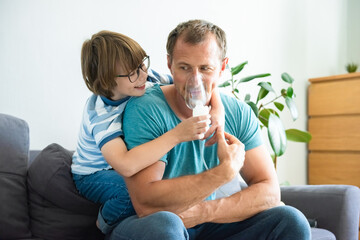 Happy child boy treats his dad at home. Medical procedures. Inhaler. Respiratory medicine. Home treatment