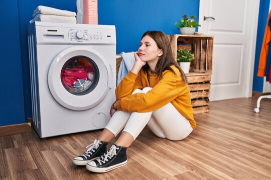 Young blonde woman sitting on floor waiting for washing machine at laundry room