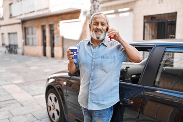 Senior grey-haired man talking on smartphone drinking coffee standing by car at street