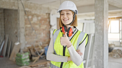 Young blonde woman architect standing with arms crossed gesture doing thumb up gesture at construction site