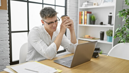 Fototapeta premium Handsome young caucasian man, confident worker smiling at his desk in the office, enjoying morning espresso while working online with laptop