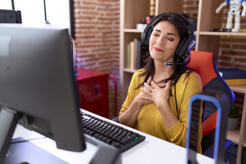 Middle age hispanic woman playing video games using headphones smiling with hands on chest with closed eyes and grateful gesture on face. health concept.