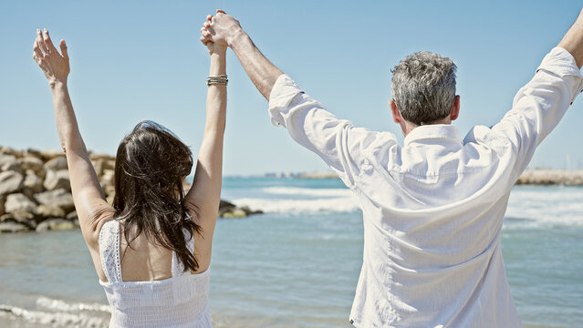 Senior man and woman couple standing with hands together and arms raised up at seaside