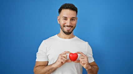 Young arab man smiling confident holding heart over isolated blue background