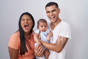 Young hispanic couple with baby standing together over isolated background angry and mad screaming...