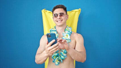 Young hispanic man tourist using smartphone relaxed on inflatable pool hammock over isolated blue...
