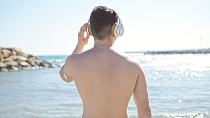 Young hispanic man tourist listening to music standing backwards at seaside