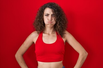 Hispanic woman with curly hair standing over red background puffing cheeks with funny face. mouth inflated with air, crazy expression. - obrazy, fototapety, plakaty