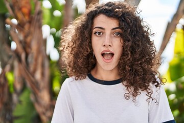 Hispanic woman with curly hair standing outdoors scared and amazed with open mouth for surprise,...