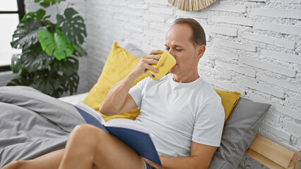 Handsome middle age man, comfortably sitting in his cozy bedroom, deeply engrossed in reading a...
