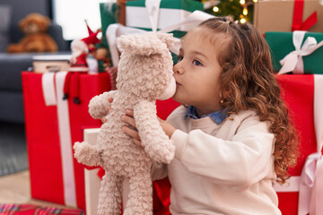 Adorable blonde toddler kissing teddy bear sitting on floor by christmas tree at home