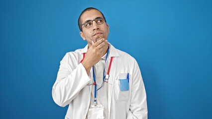 Young hispanic man doctor standing with doubt expression thinking over isolated blue background