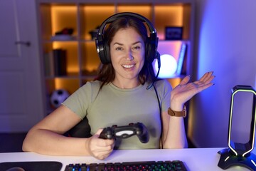 Beautiful brunette woman playing video games wearing headphones pointing aside with hands open palms showing copy space, presenting advertisement smiling excited happy