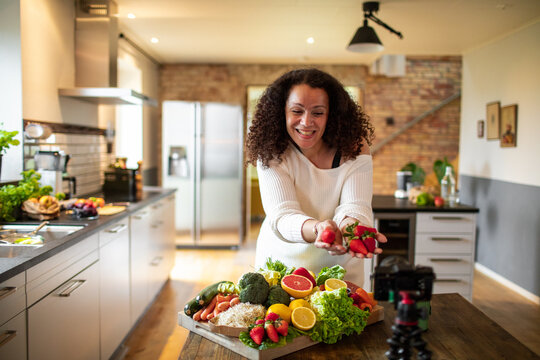 Middle aged female mixed ethnicity food nutritionist and blogger recording a video about a healthy recipe with organic fruits and vegetables in her kitchen at home