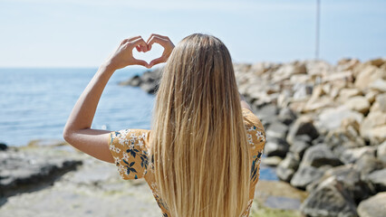 Fototapeta na wymiar Young blonde woman doing heart gesture backwards at seaside