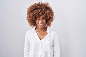 Young hispanic woman with curly hair standing over white background winking looking at the camera with sexy expression, cheerful and happy face.