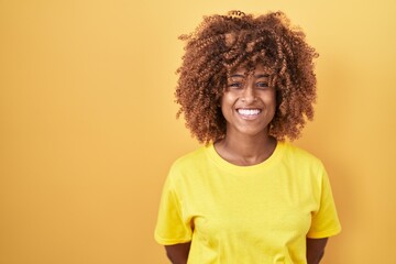 Young hispanic woman with curly hair standing over yellow background with a happy and cool smile on face. lucky person.