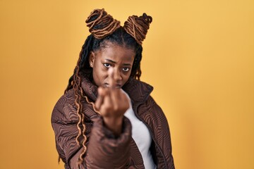 African woman with braided hair standing over yellow background showing middle finger, impolite and rude fuck off expression