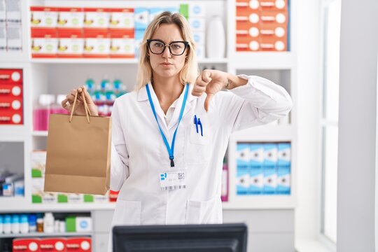 Young blonde woman working at pharmacy drugstore holding paper bag with angry face, negative sign showing dislike with thumbs down, rejection concept