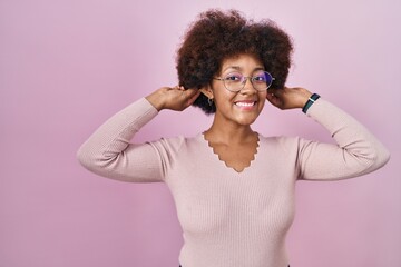 Young african american woman standing over pink background smiling pulling ears with fingers, funny gesture. audition problem