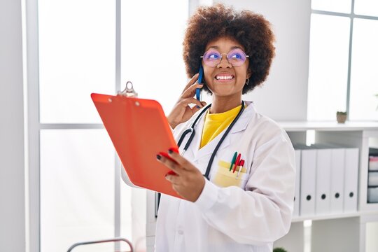 African american woman wearing doctor uniform talking on smartphone reading document at clinic