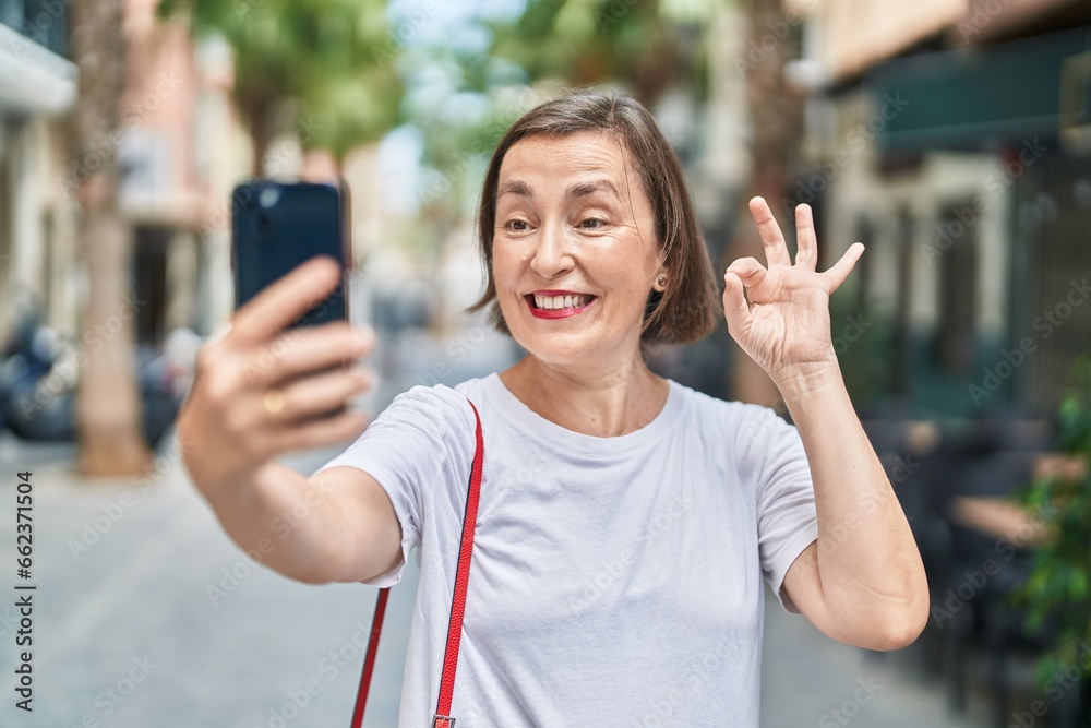 Wall mural Middle age hispanic woman doing video call with smartphone doing ok sign with fingers, smiling friendly gesturing excellent symbol