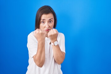 Middle age hispanic woman standing over blue background ready to fight with fist defense gesture, angry and upset face, afraid of problem