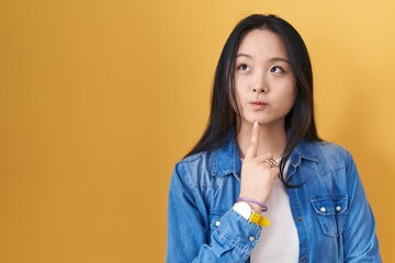 Young asian woman standing over yellow background thinking concentrated about doubt with finger on...
