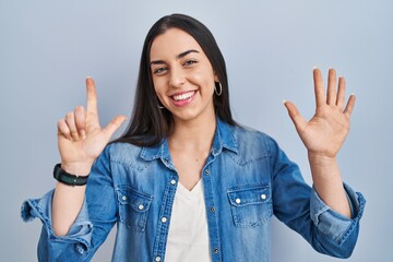 Hispanic woman standing over blue background showing and pointing up with fingers number seven while smiling confident and happy.
