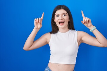 Fototapeta premium Young caucasian woman standing over blue background smiling amazed and surprised and pointing up with fingers and raised arms.