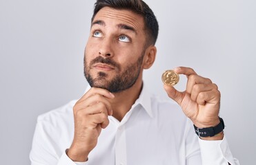 Handsome hispanic man holding litecoin cryptocurrency coin serious face thinking about question with hand on chin, thoughtful about confusing idea